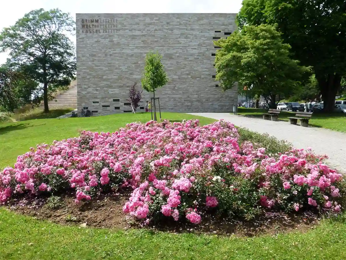 Der helle Steinbau der Grimmwelt Kassel mit Schriftzug, davor ein Rosenbeet und Bäume im Sommer.